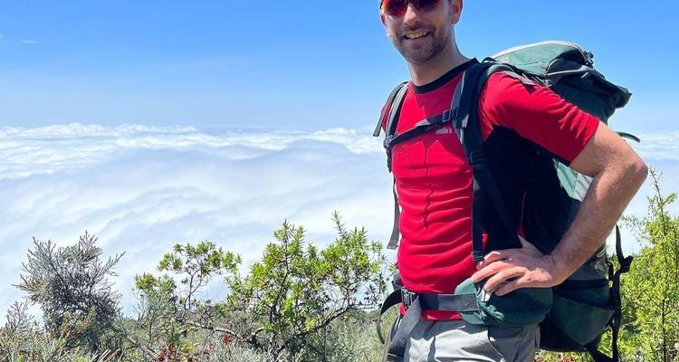 Ein Mann mit einem roten Hemd und Rucksack, der auf einem Berg mit Wolken darunter steht.