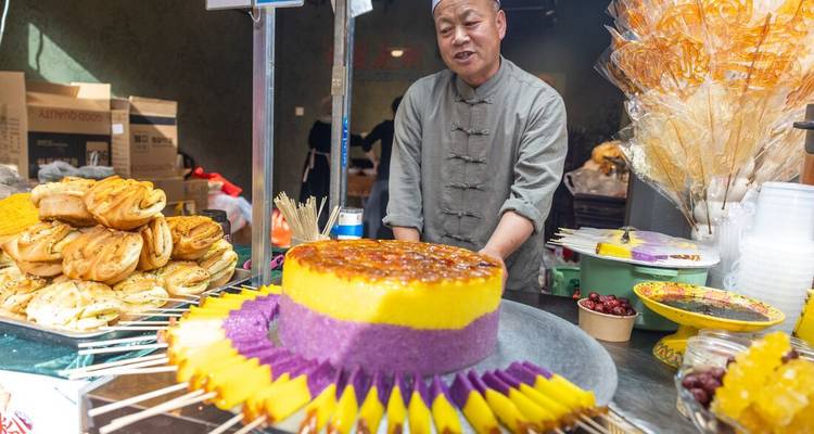 Un vendeur de rue présente fièrement un gâteau de riz violet et jaune vibrant entouré d'échantillons sur brochettes à son étal.