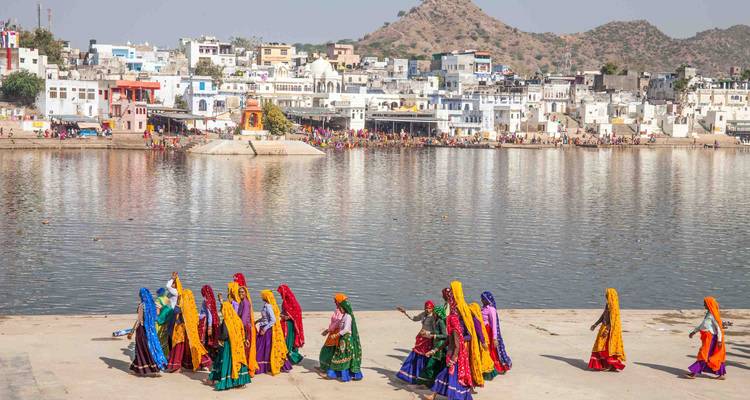 Frauen in leuchtenden traditionellen Saris gehen am Pushkar Lake entlang, während sich die weiß getünchte Stadt und karge Hügel dahinter erheben.
