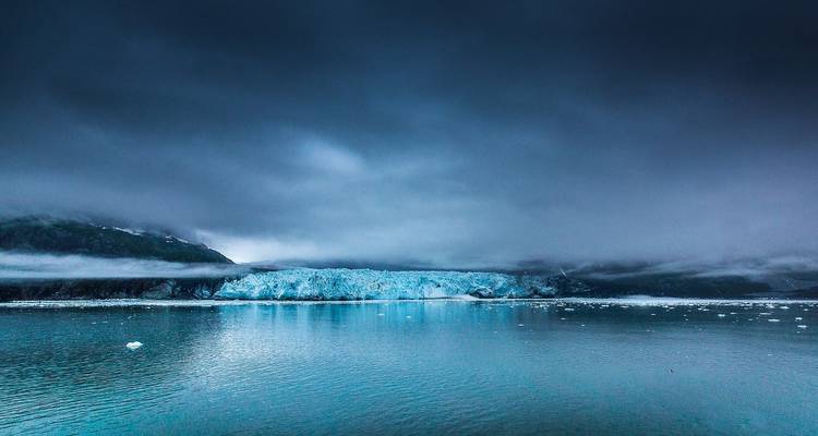 Moody seascape with a wide blue glacier meeting calm grey water under low clouds and mist.