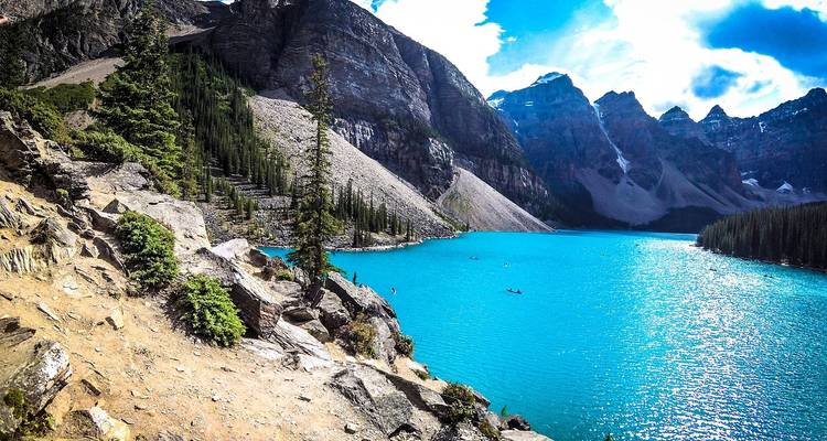 Brilliant turquoise Moraine Lake surrounded by rugged peaks and pine forest, hikers on rocky shore.