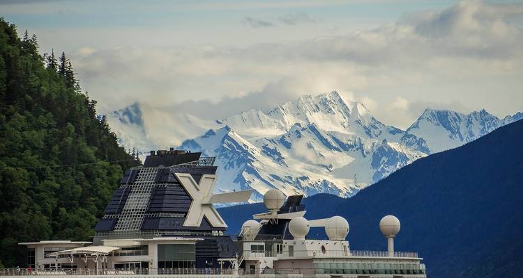 Large modern cruise ship sails past verdant hills toward snow-covered mountain range under cloudy sky.