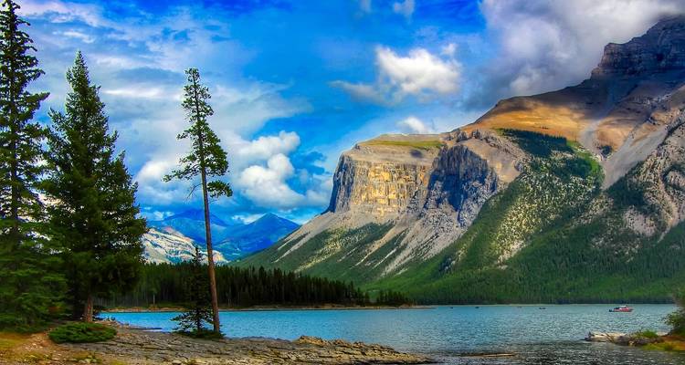 Dramatic cliff face and pine trees rise above a tranquil mountain lake under patchy clouds.
