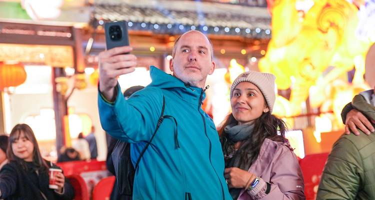 Pareja tomándose una selfie con el smartphone rodeada de decoraciones festivas iluminadas y coloridas por la noche