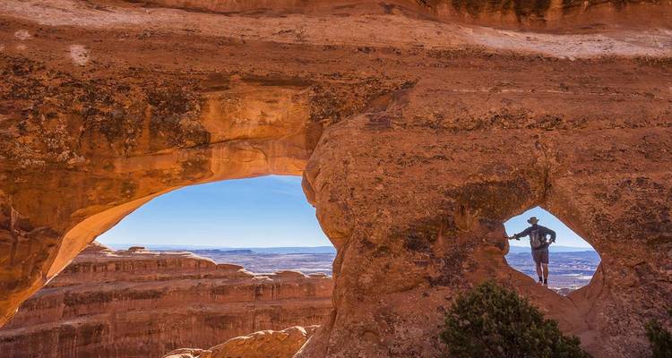 Excursionista enmarcado por una ventana natural de arenisca con vista a vastos cañones de roca roja bajo un cielo brillante.