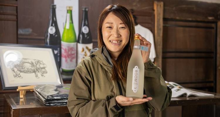Femme souriante qui présente fièrement une bouteille de saké dans une boutique traditionnelle de brasserie japonaise.