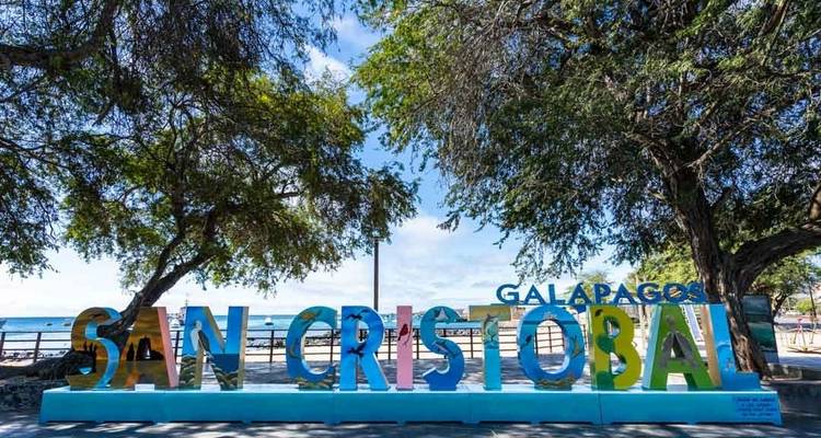 Colorful sign reading 'San Cristobal' with trees and ocean in the background.