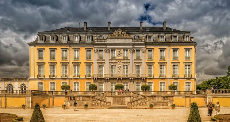 Baroque yellow palace stands under dramatic cloudy skies with visitors in the courtyard.