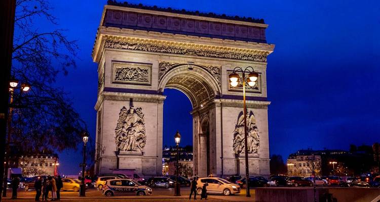 Arc de Triomphe illuminated at night with vehicles and pedestrians passing beneath a deep blue sky.