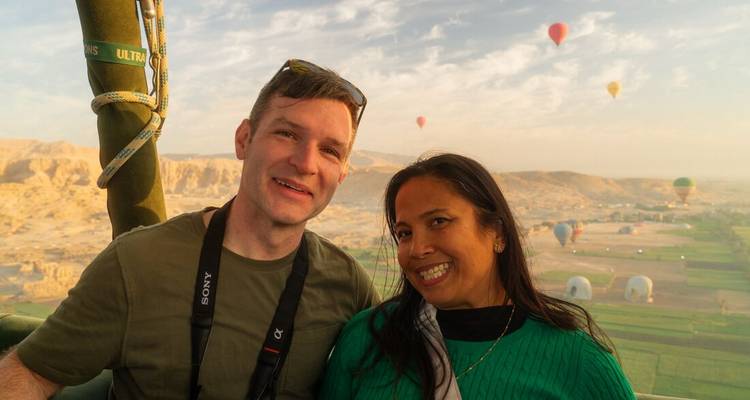 Pareja sonriente en una canasta de globo aerostático flotando sobre campos verdes y colinas desérticas salpicadas de globos coloridos en la suave luz matutina sobre Luxor.