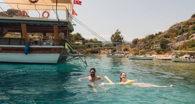 Un hombre y una mujer flotan juguetonamente en agua turquesa cristalina junto a un barco de madera tradicional que ondea la bandera turca.