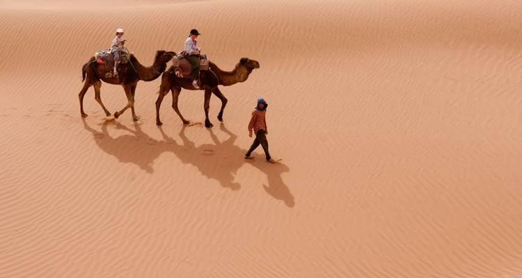 Zwei Personen, die auf Kamelen reiten, geführt von einem Führer in einer Wüste.