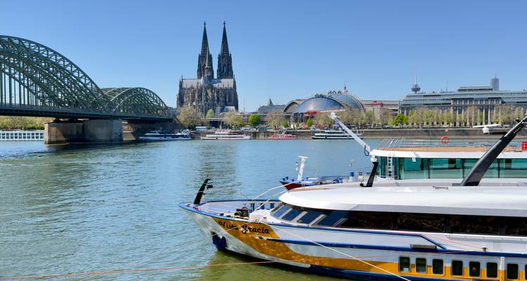 Rhine riverboat in front of Cologne Cathedral and arched railway bridge on a clear day.