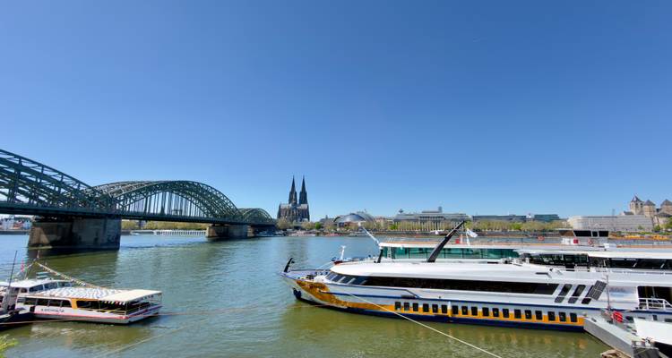 Wide angled view of Cologne waterfront with cathedral, bridge and several moored vessels.