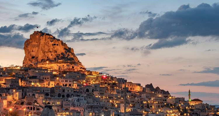 Uçhisar Castle and surrounding cave houses of Cappadocia glow with warm lights under a pastel evening sky