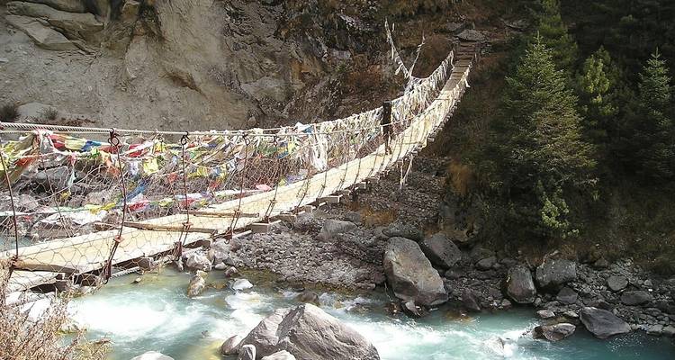 Puente colgante adornado con banderas de oración se extiende sobre un cañón de río himalayo turquesa.