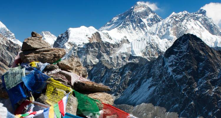 Coloridas banderas de oración cuelgan sobre un montículo de piedras enmarcando una vista dramática del Monte Everest y los picos nevados circundantes.