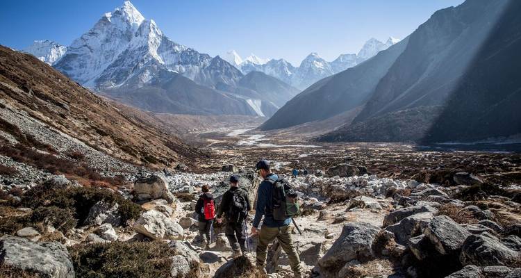 Los excursionistas descienden por un sendero rocoso hacia un dramático valle del Himalaya enmarcado por imponentes picos nevados bajo un cielo despejado.