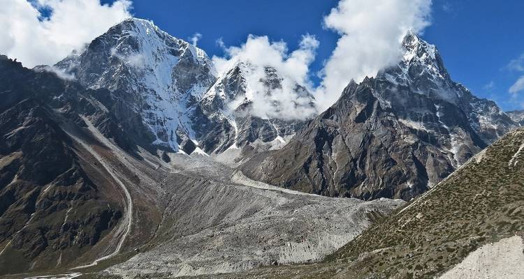 Picos himalayos dentados coronados de nieve se elevan abruptamente sobre morrenas rocosas y nubes tenues.