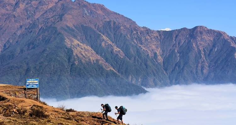 Two hikers walking on a hill with clouds below and mountains in the background.