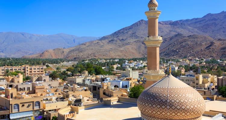 Gouden minaret en koepelmoskee die uitsteken boven het adobestadje Nizwa met bergen op de achtergrond.