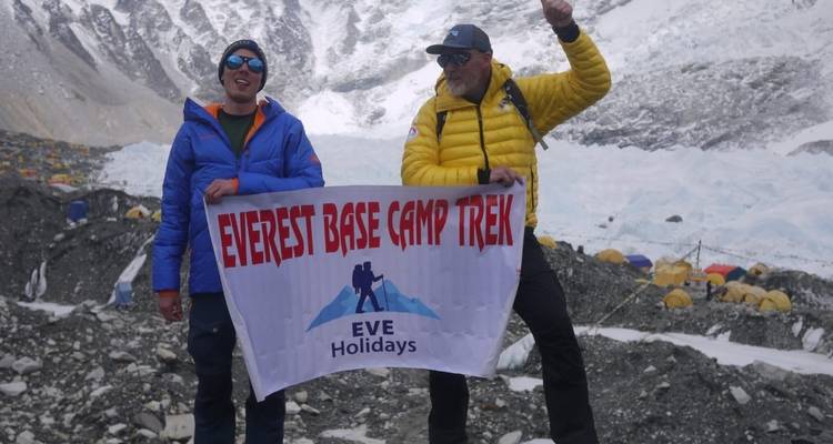 Two people holding a banner for Everest Base Camp Trek with tents and mountains in the background.