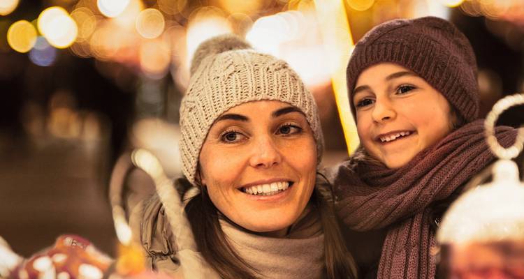 Madre e hijo abrigados con gorros de invierno sonriendo entre luces navideñas brillantes.