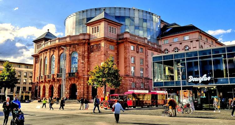 Edificio de teatro moderno con techo de cristal en una plaza animada en Maguncia con peatones y un pequeño tren turístico.