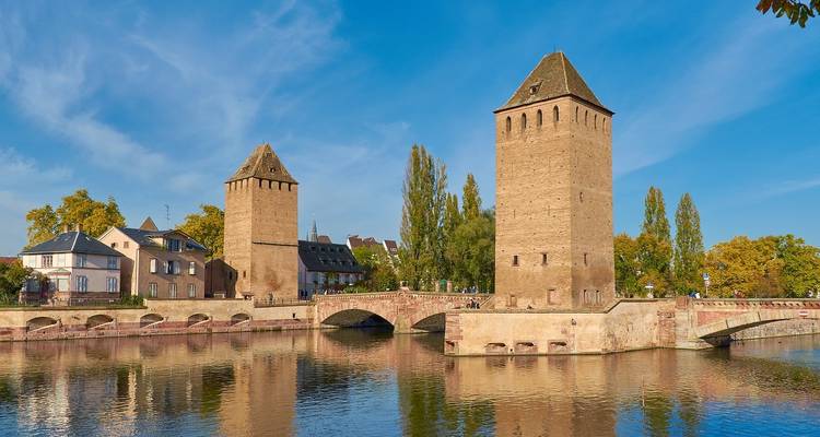 Torres históricas de piedra y puente reflejados en el río Ill bajo un cielo azul despejado en Estrasburgo.