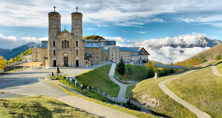 Sanctuaire de montagne et basilique perchés sur un plateau alpin avec des nuages tourbillonnant autour de pics lointains.