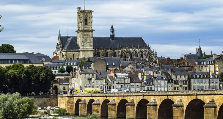 Ville française historique avec une église haute et des toits en ardoise, vue à travers un ancien pont en pierre.
