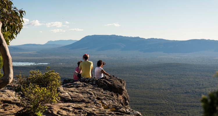 Drie reizigers zitten op een rotsachtige kliffrand en kijken uit over uitgestrekte beboste bergketens in de Grampians.