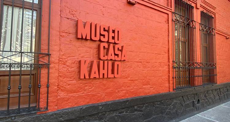 Pared naranja brillante con letrero llamativo que dice 'Museo Casa Kahlo' junto a ventanas de hierro forjado.