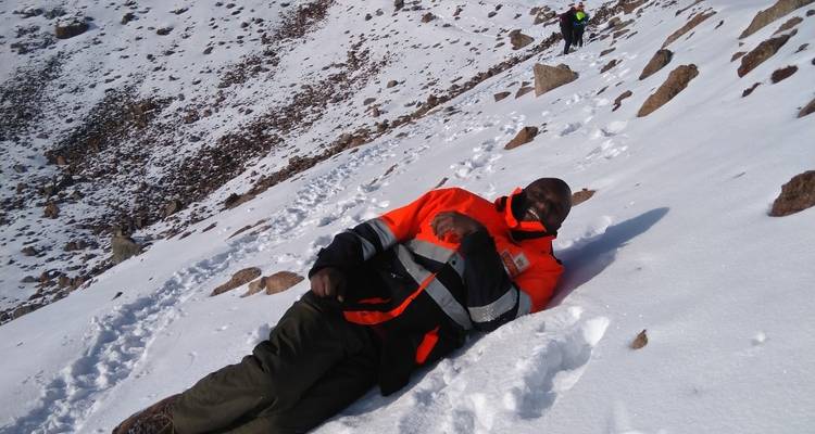 A person in an orange jacket lying on a snowy mountainside with another person in the background.