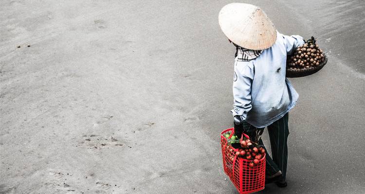 Un vendedor callejero con un sombrero cónico lleva canastas de fruta fresca por una calle gris urbana.