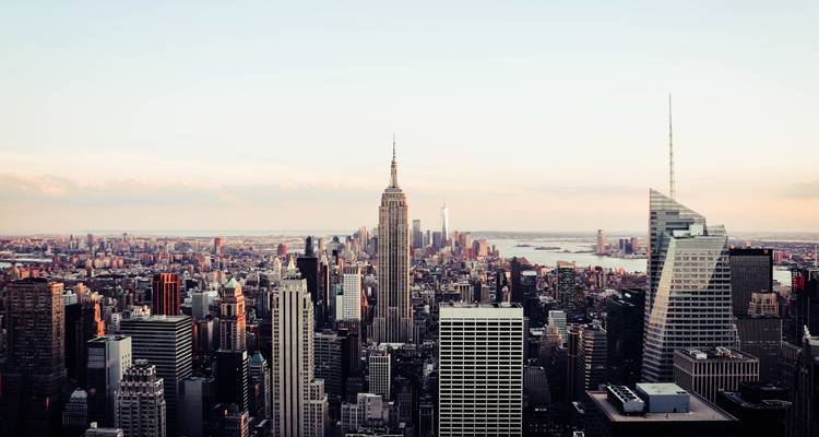 Vue panoramique large de New York avec le Empire State Building s'élevant au-dessus des gratte-ciel environnants sous un ciel de soirée aux tons pastel.