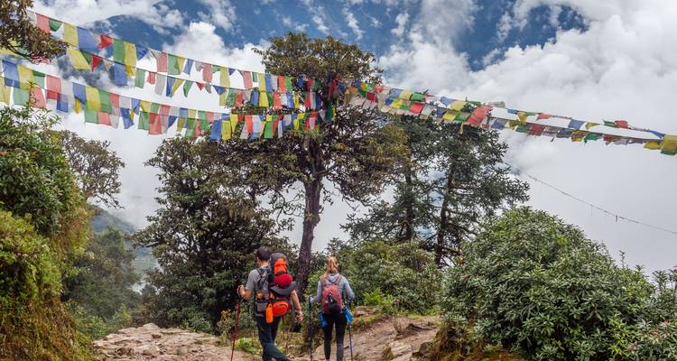 Randonneurs sur un sentier avec des drapeaux de prière et des nuages en contrebas.