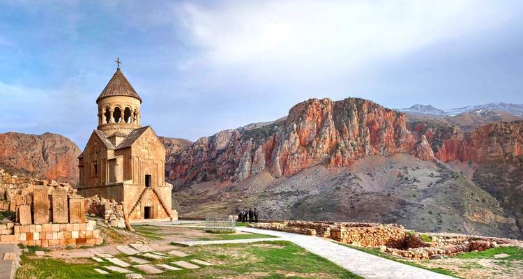 Monastery with red stone structure against dramatic cliff backdrop.