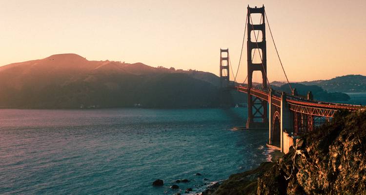 The Golden Gate Bridge stretches across the mouth of San Francisco Bay bathed in warm sunset light with rolling hills beyond.