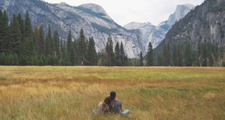 A couple sits in a golden meadow beneath towering granite cliffs and pine forests in Yosemite National Park.
