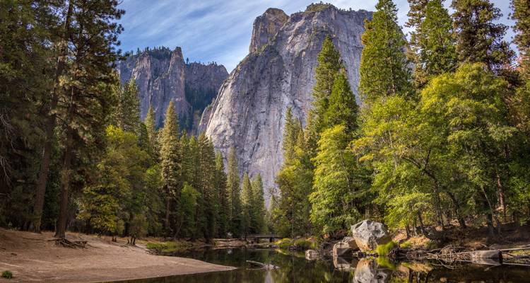 Riverside reflections frame soaring granite cliffs and dense pine forest in Yosemite Valley on a crisp day.