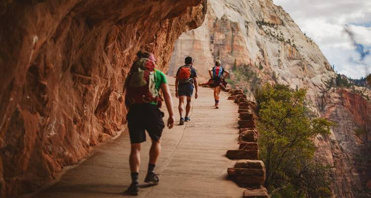 Excursionistas con mochilas atraviesan un sendero estrecho al borde del acantilado en las paredes de arenisca roja del Parque Nacional Zion.