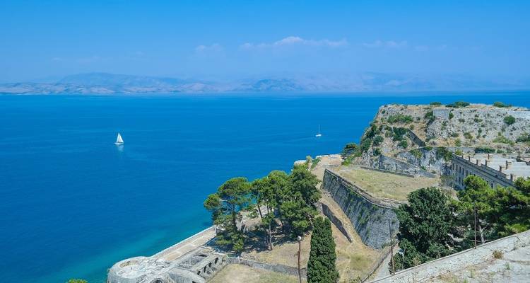 Sunny coastal panorama of Corfu’s old fortress walls jutting into vivid blue Ionian Sea with distant hazy mountains.