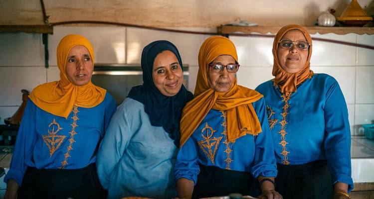Vier Marokkaanse vrouwen in bijpassende blauwe en mosterdgele outfits glimlachen in een keukenomgeving.