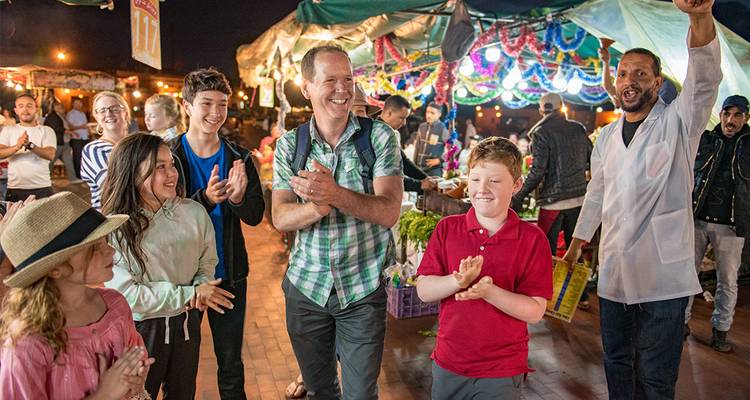 Nachtelijke marktscène met families die meeklappen langs een levendige straat vol kraampjes in Marrakesh's Jemaa el-Fna.