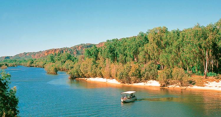 Petit bateau de croisière naviguant le long d'une rivière calme bordée d'une dense forêt d'eucalyptus sous un ciel bleu dégagé.