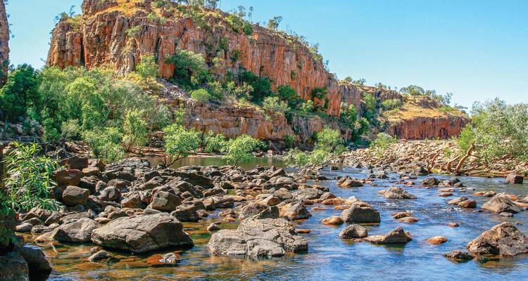 Felsiger Fluss mit Felsbrocken, der unter orangefarbenen Sandsteinklippen in Australiens Outback fließt.