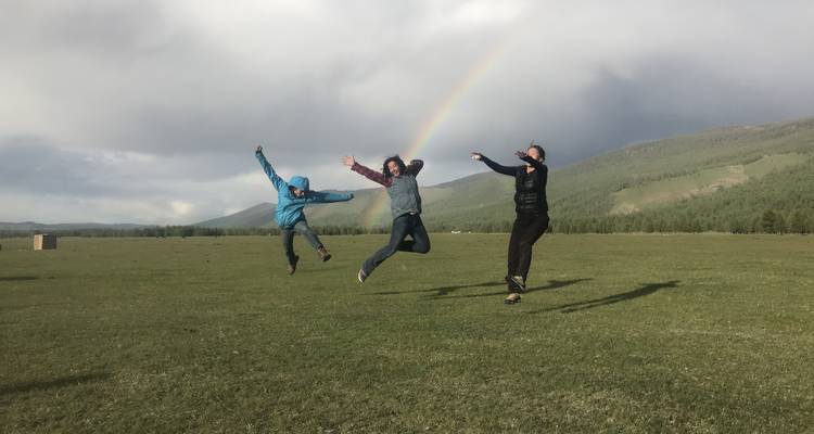 Drei Menschen springen fröhlich auf einem Feld mit einem Regenbogen im Hintergrund.