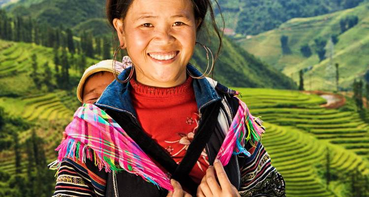 Femme souriante en tenue traditionnelle colorée porte un bébé avec des rizières en terrasses derrière.