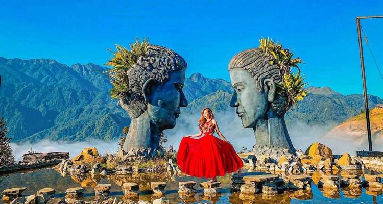 Femme en robe rouge fluide assise entre deux grandes sculptures de visages avec des montagnes brumeuses derrière à Sapa.